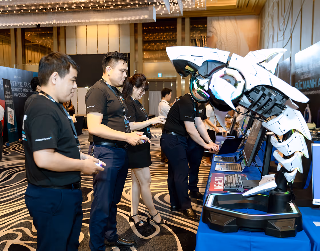Three men sitting at a table with bottled water and flowers, attentively watching and clapping during an indoor event with a large audience.