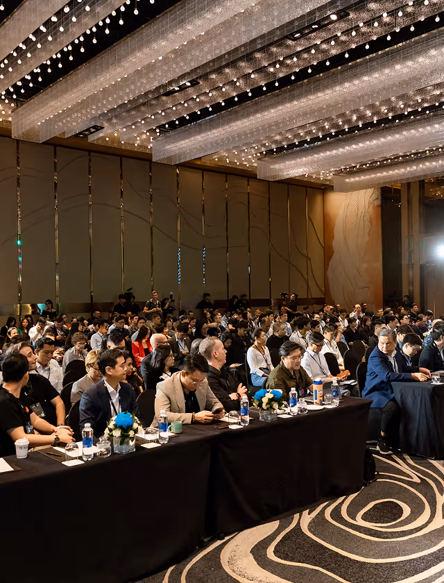 Large audience seated in rows at a conference or seminar in a modern hall with decorative ceiling lights.