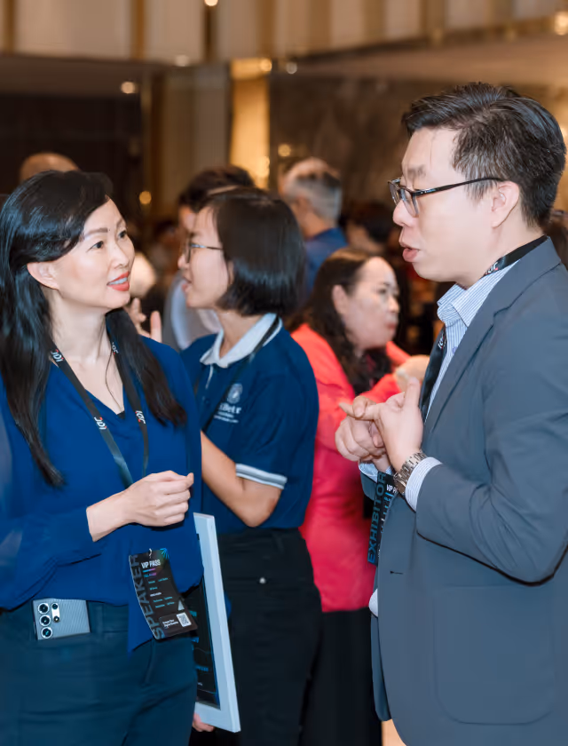 A man in glasses and a suit chatting with a woman in a blue blouse at a conference, with other attendees in the background.