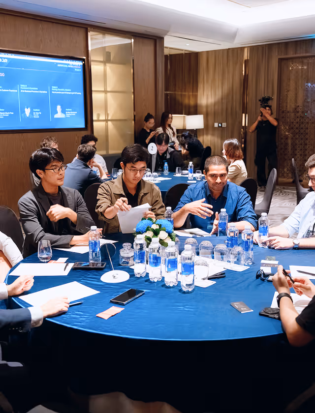 Group of professionals seated around a round table with water bottles and documents, engaging in a meeting or discussion in a modern conference room.