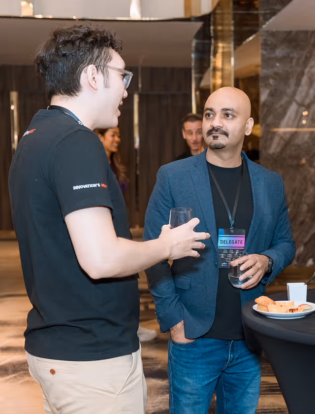 Two men conversing at a networking event, one wearing a delegate badge and holding a glass, standing near a table with snacks and a drink.