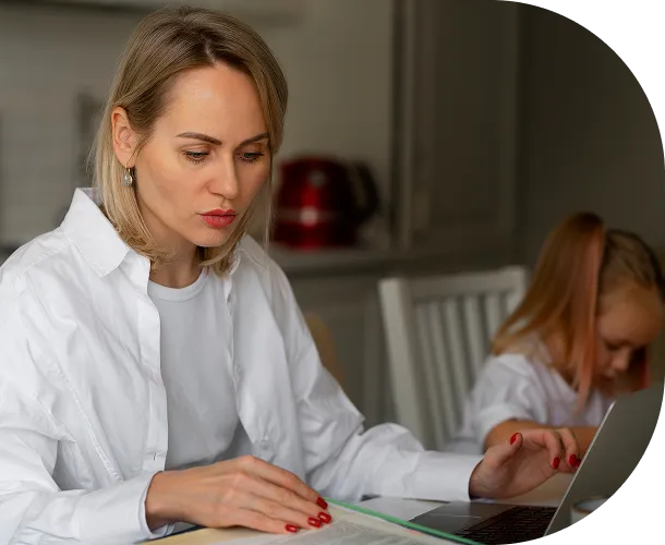 Woman in white shirt working on a laptop at a table with a small girl sitting next to her.