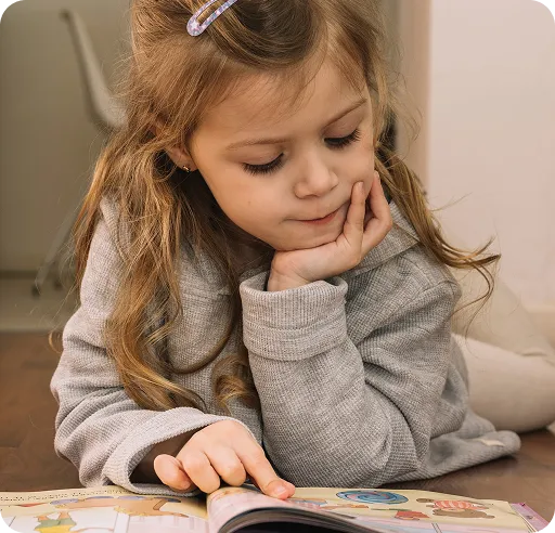 Young girl with long hair and a hair clip, wearing a grey sweater, lying on the floor and reading a book.