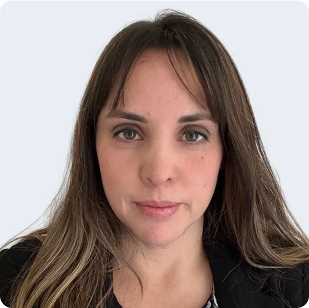 Portrait of a woman with light skin, long brown hair with highlights, and green eyes wearing a black jacket over a patterned top against a plain light background.