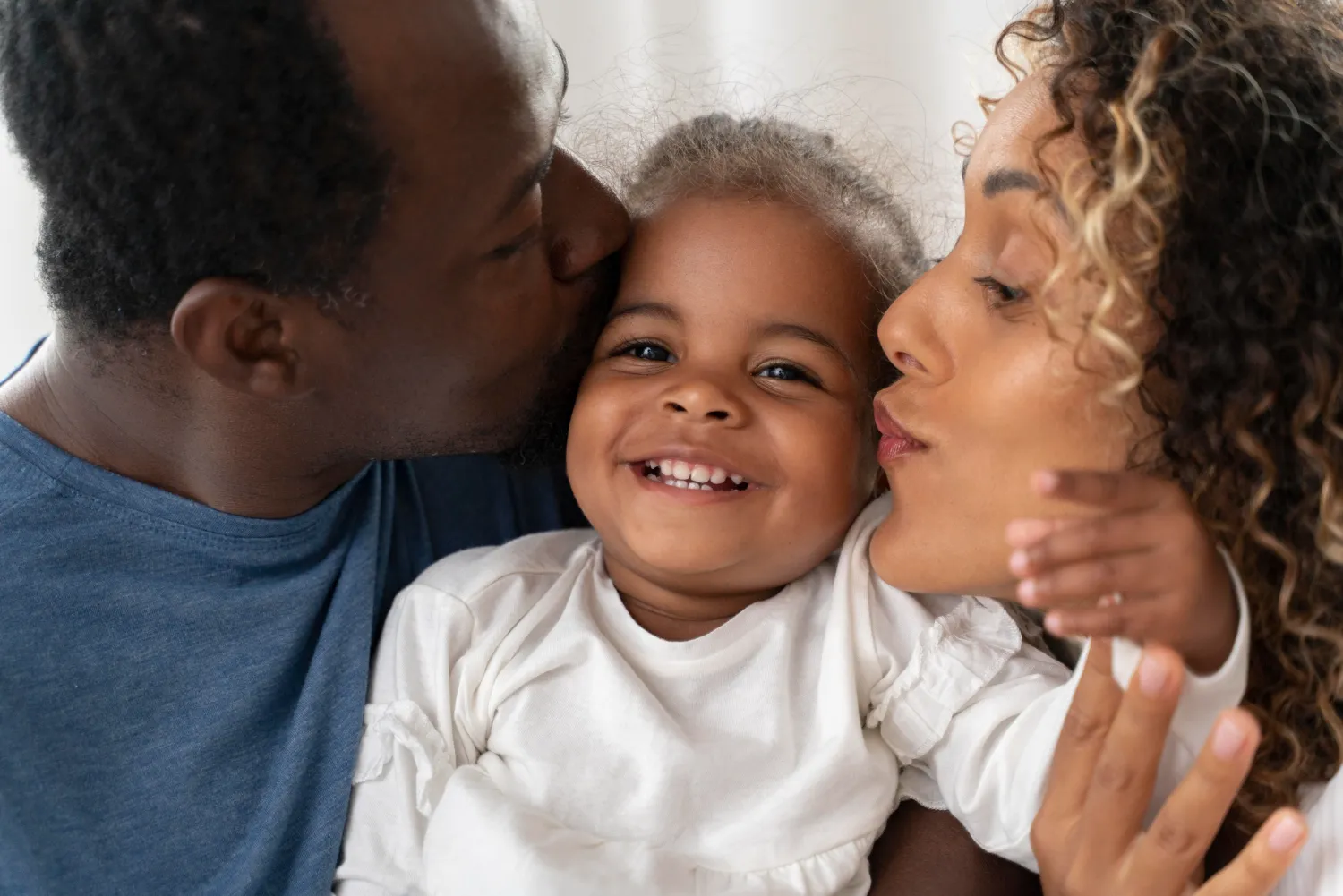 Smiling toddler being kissed on both cheeks by a man and a woman with curly hair.