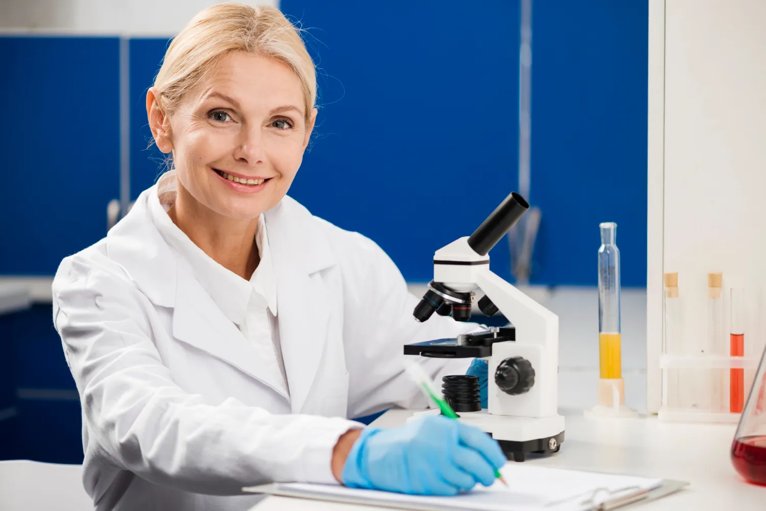 Smiling female scientist in a white lab coat and blue gloves working with a microscope and writing notes in a laboratory.