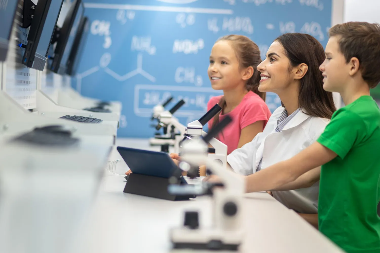 A female teacher and two children in a science classroom looking at computer screens with microscopes on the table.