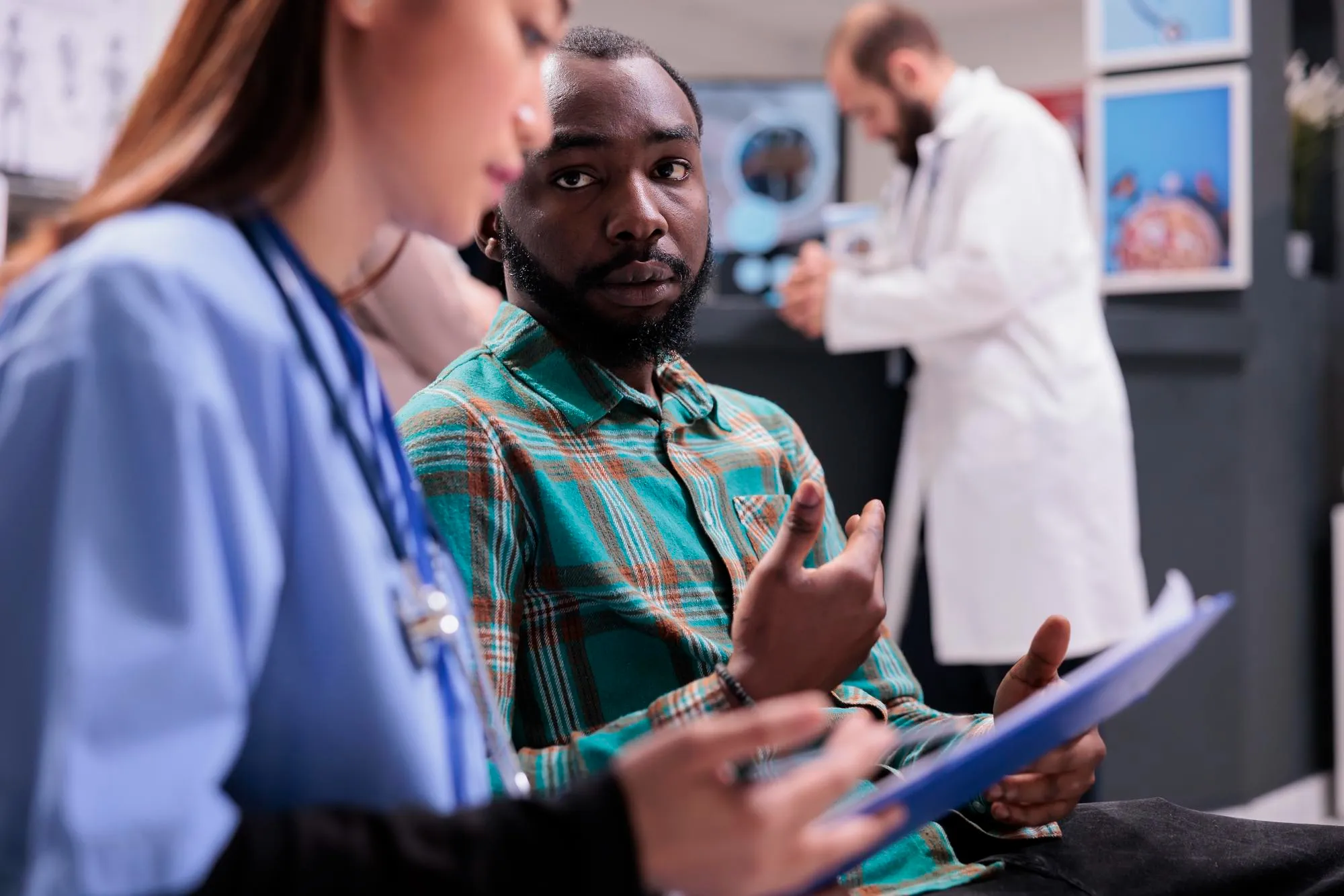Man in green plaid shirt discussing medical information with a female healthcare professional holding a clipboard, with a doctor in a white coat in the background.