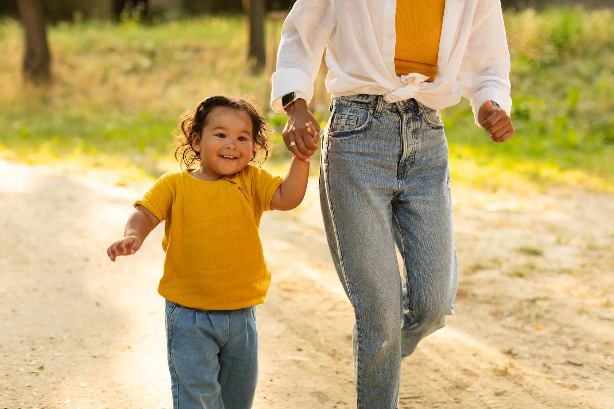 Smiling toddler wearing a yellow shirt walking hand-in-hand with an adult in blue jeans on a sunlit path.