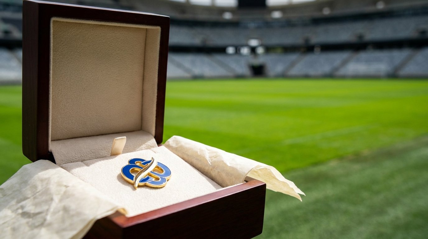 Geöffnete Merchandise-Geschenkbox mit Emaille-Pin und Seidenpapier am Spielfeldrand, dahinter unscharf leere Tribünen