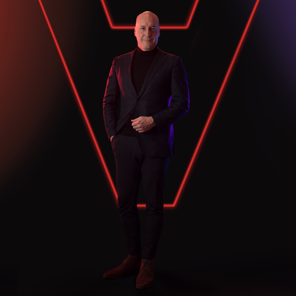 Portrait of a bald man wearing a black turtleneck and dark blazer against a black background with red and purple lighting.
