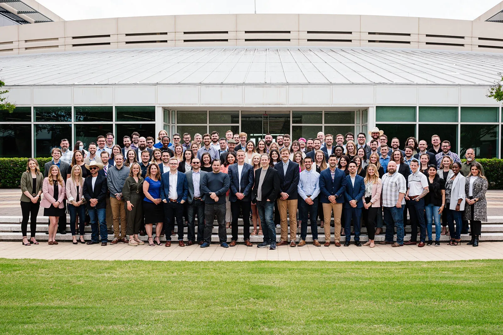 Leadr group photo of diverse employees standing in front of a modern office building with large windows and green lawn.