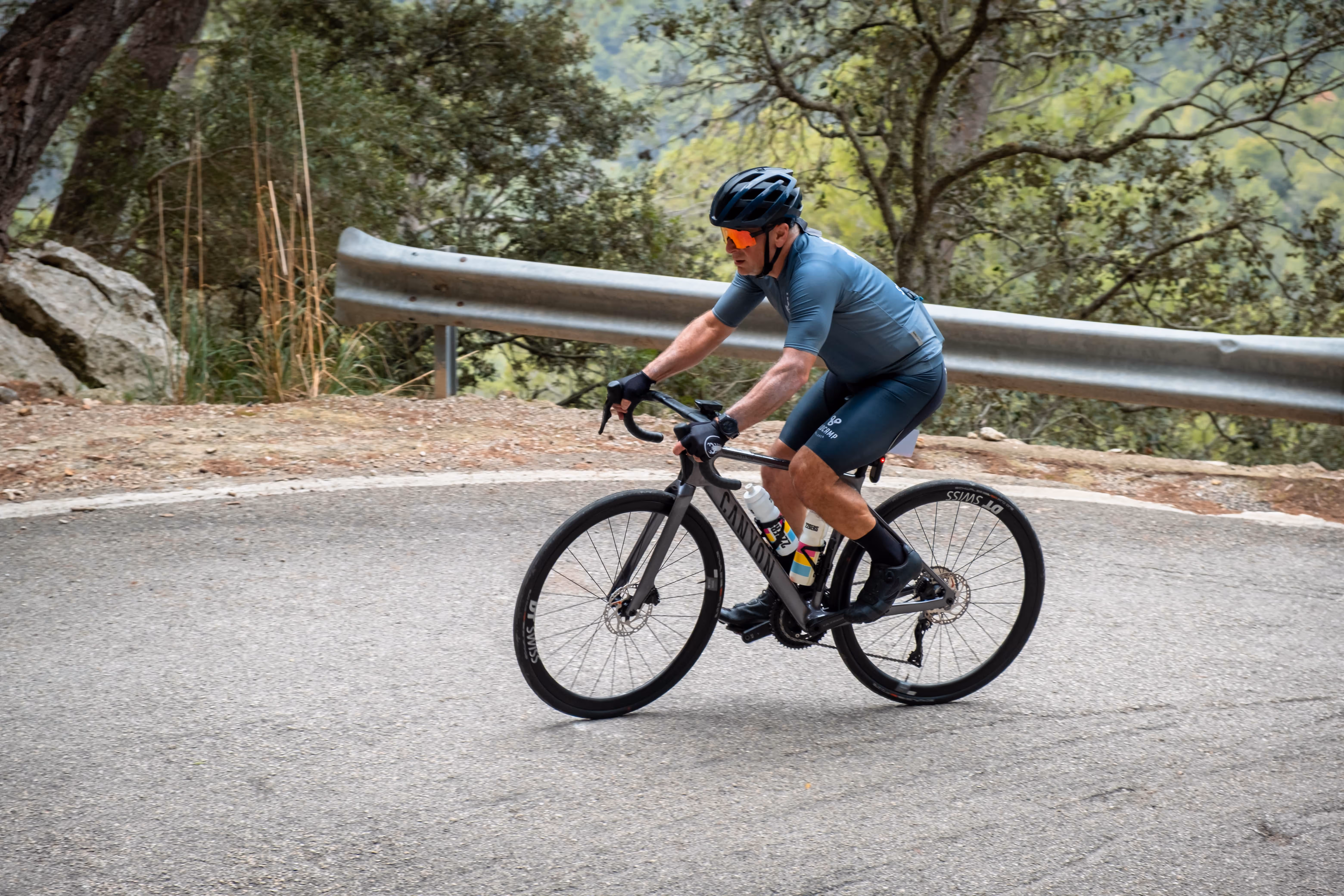 Cyclist in blue cycling gear riding a Canyon road bike on a curved mountain road with a metal guardrail and trees in the background.