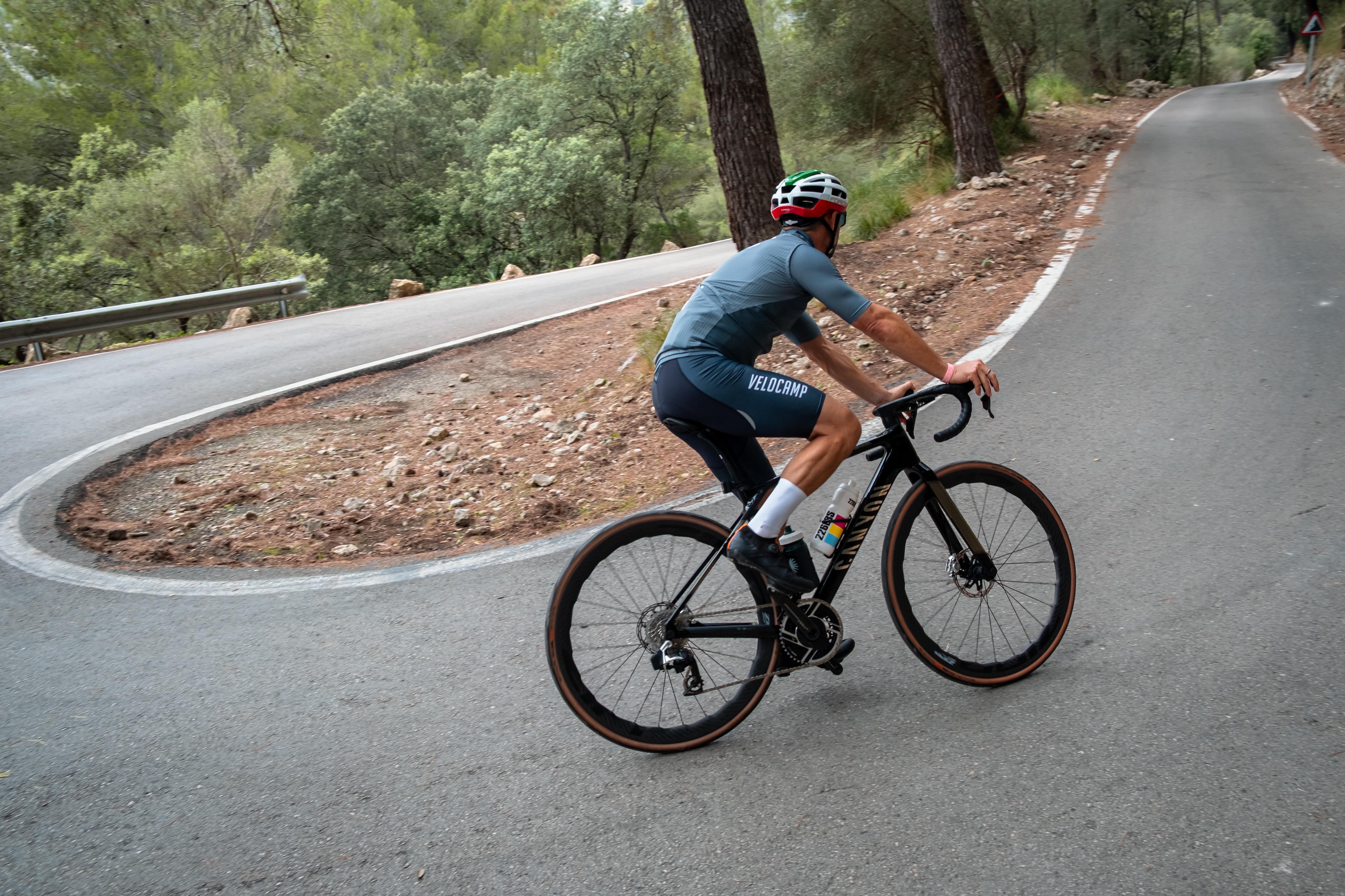 Cyclist wearing a helmet and blue cycling outfit riding a black road bike on a winding mountain road surrounded by trees.