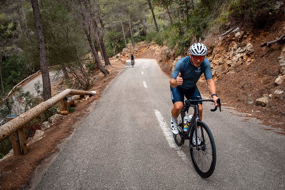 Cyclist in blue gear and white helmet giving thumbs up while riding on a winding forest road with another cyclist in the distance.