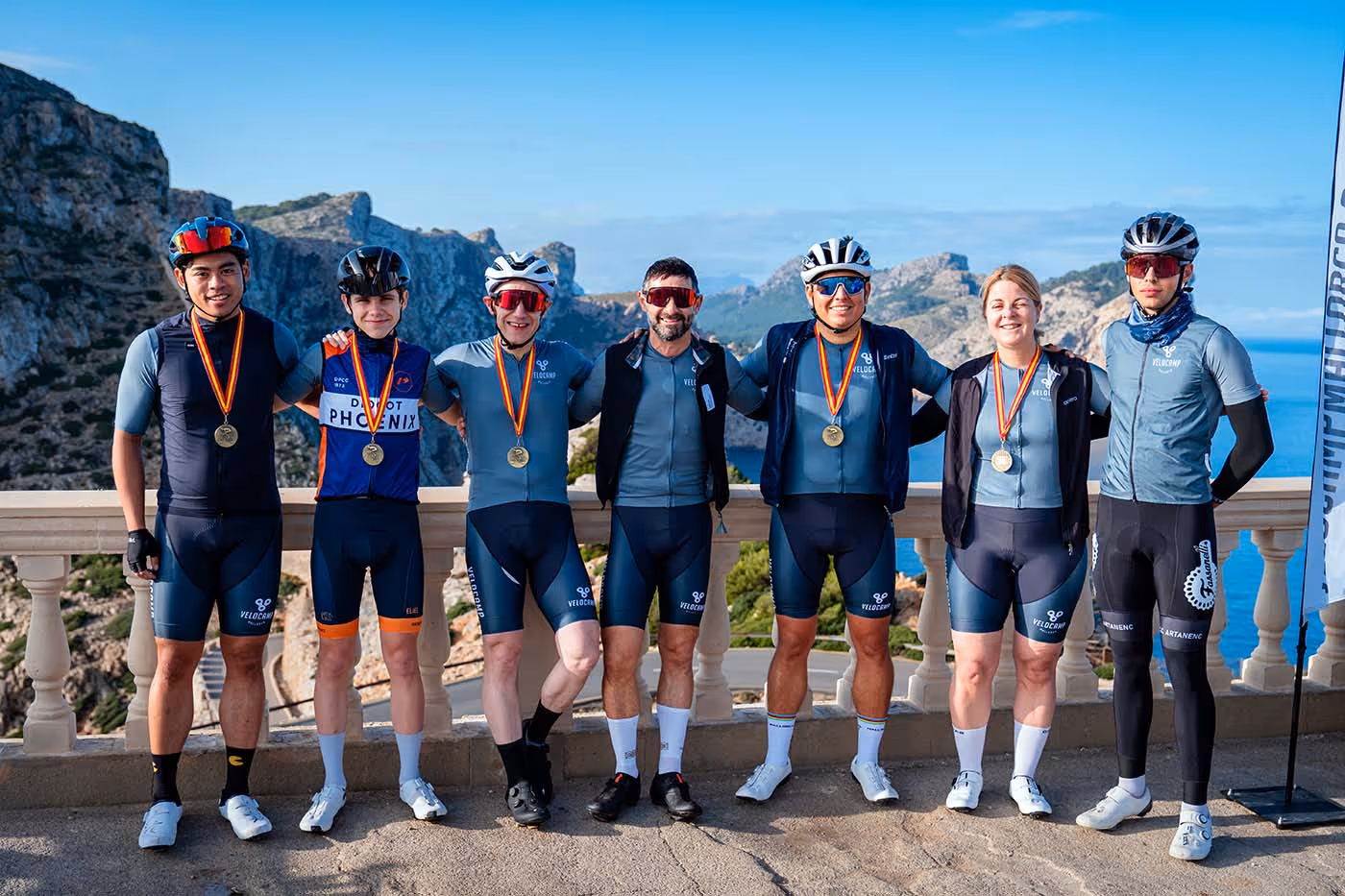 Seven cyclists wearing medals stand side by side outdoors with mountainous landscape and blue sky in the background.