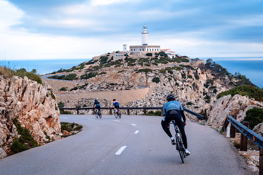 Three cyclists riding on a winding road along rocky cliffs towards a lighthouse on a hill under a cloudy sky.