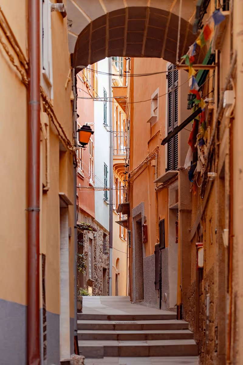Narrow alley with stone steps and orange and beige buildings with shuttered windows and an arch overhead.