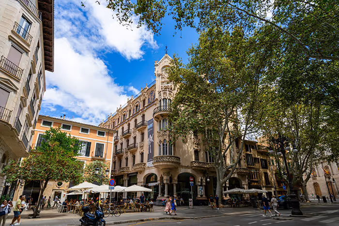 Urban street scene with historic buildings, outdoor café seating, and pedestrians under a partly cloudy blue sky.