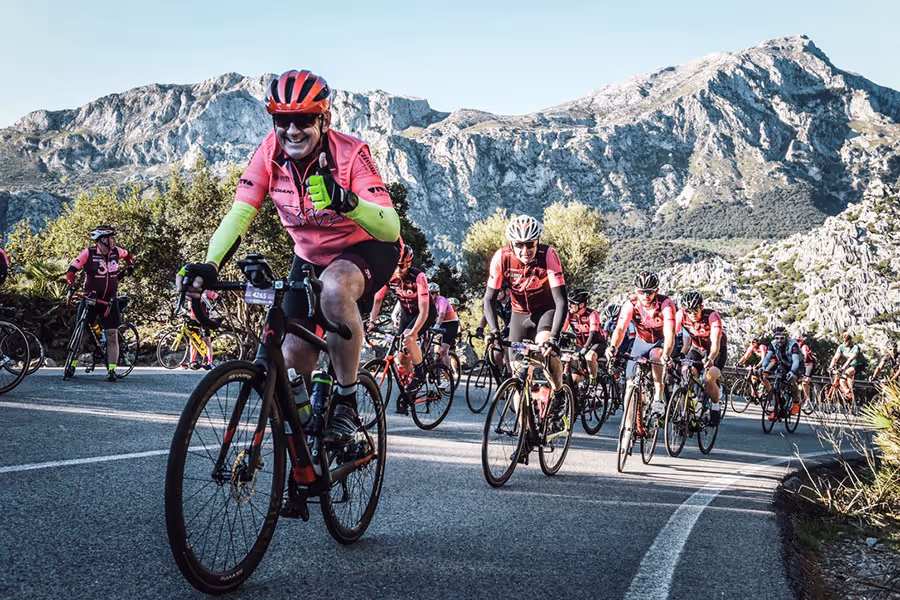 Group of cyclists wearing pink and black gear riding on a winding road with mountainous landscape in the background, one cyclist in front giving a thumbs-up.