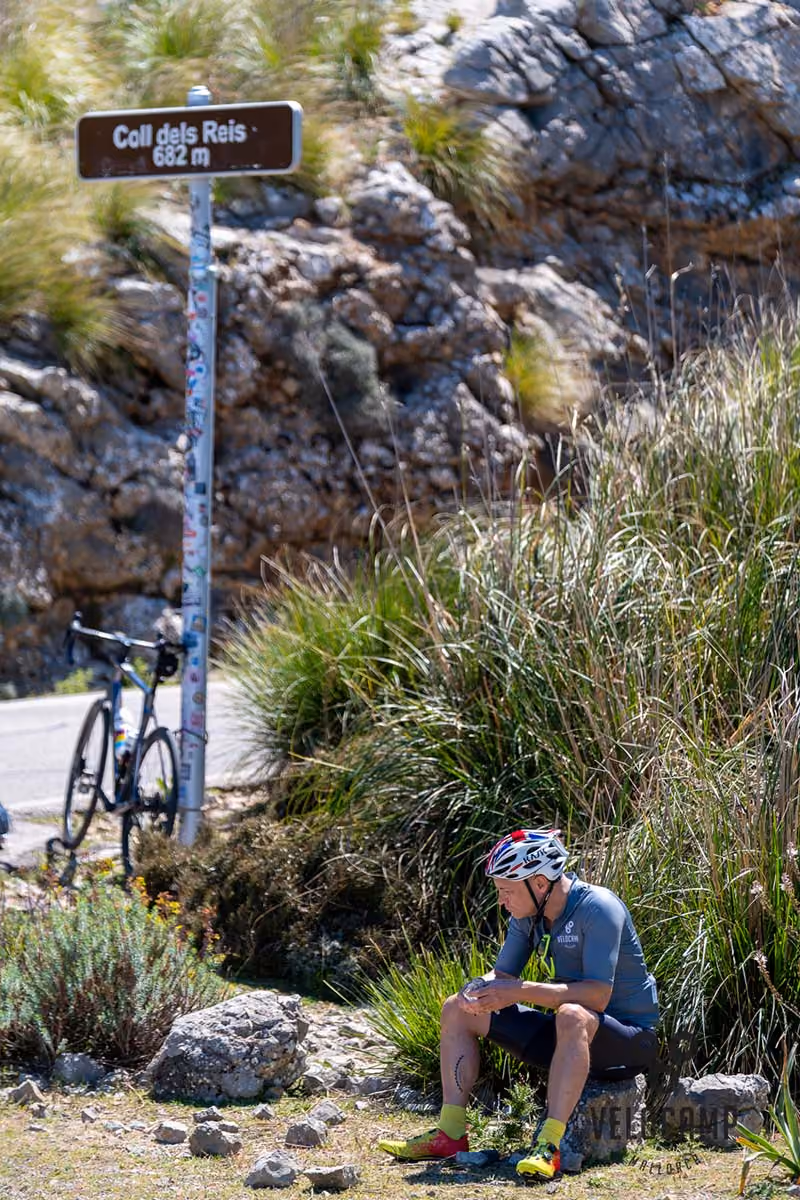 Cyclist in helmet and gear sitting on rock near a sign reading Coll dels Reis 682 m in rocky landscape.