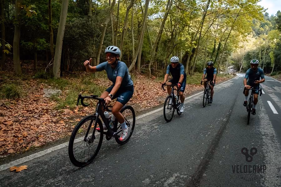 Four cyclists riding on a forested road, one smiling and giving a thumbs-up gesture.