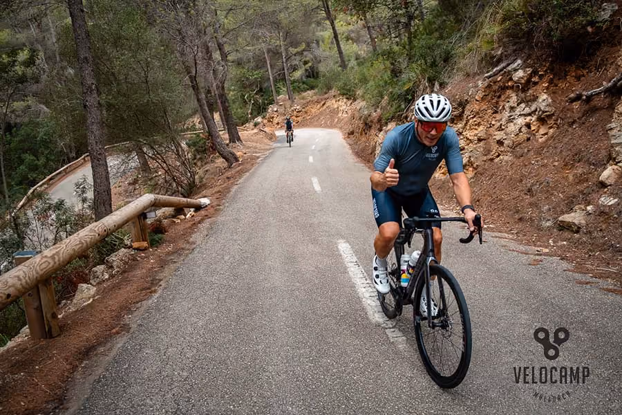 Cyclist in blue gear giving thumbs up while riding on a mountain road surrounded by trees.