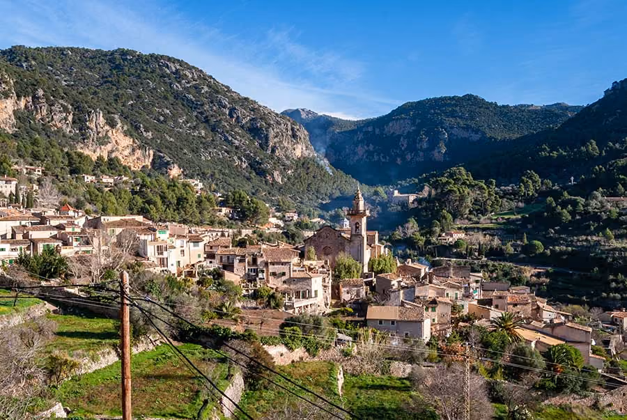 Small village with a church tower nestled in a green valley surrounded by mountains under a blue sky.