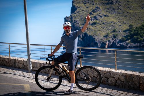 Cyclist wearing a white helmet, red sunglasses, and gray cycling outfit sitting on a black Canyon road bike with two gold water bottles, raising his right arm with one finger pointing up on a sunny seaside road with rocky cliffs and calm water in the background.