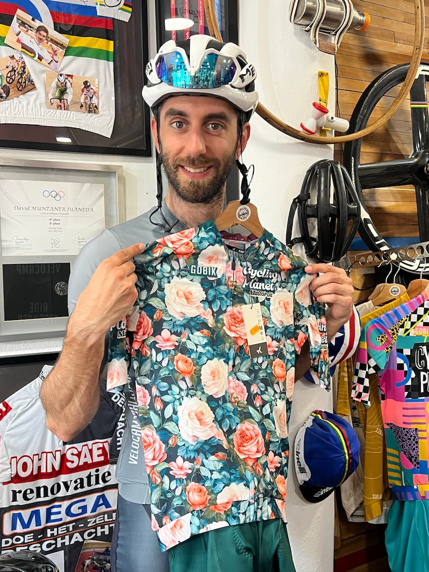 Man wearing a white cycling helmet and mirrored sunglasses holds up a floral cycling jersey with 'GOBIK' and 'Cycling Planet Mallorca' logos inside a shop with cycling memorabilia and colorful jerseys in the background.