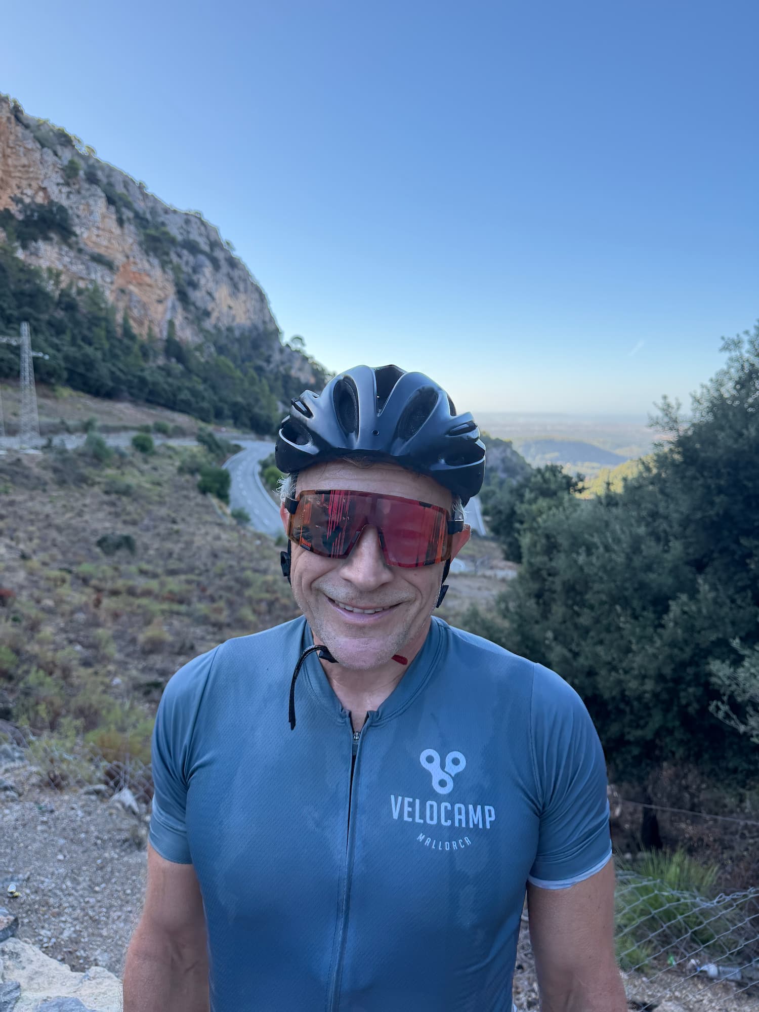 Smiling man wearing a black cycling helmet, large red sunglasses, and a blue Velocamp Mallorca cycling jersey, standing outdoors with a winding road, rocky hillside, and clear blue sky in the background.