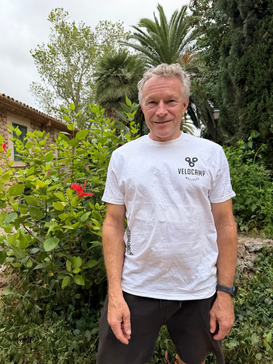 Middle-aged man with gray hair smiling, wearing a white Velocamp Mallorca t-shirt and black shorts, standing outdoors in front of green plants and trees with a stone building in the background.