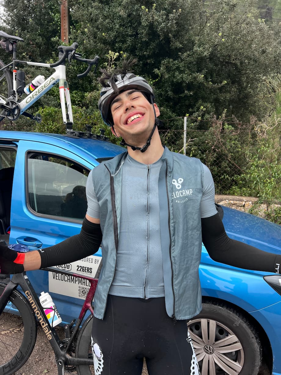 Smiling cyclist with closed eyes wearing a helmet, black arm sleeves, and a light blue sleeveless vest over a cycling jersey stands with arms open in front of a blue car with a mounted bicycle on its roof rack.