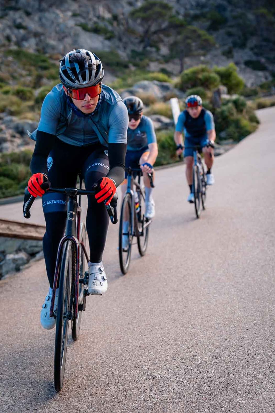 Three cyclists wearing helmets and sunglasses ride road bikes uphill on a paved path surrounded by rocky, shrub-covered terrain, with the lead cyclist wearing red gloves and white shoes.
