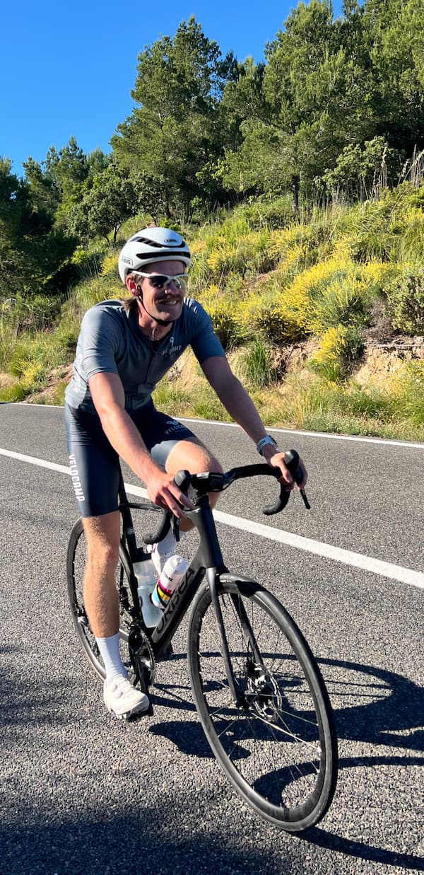 Man in gray cycling outfit and white helmet riding a black road bike on a sunny road with green trees and yellow shrubs in the background.