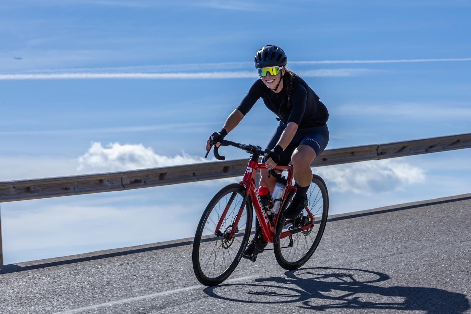 Smiling cyclist wearing a black helmet, sunglasses, and cycling gear riding a red road bike on a paved road with a wooden guardrail and blue sky with clouds in the background.