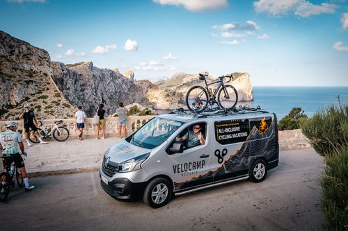 Cyclists in matching gear pause by a van on a coastal mountain road under clear blue sky.