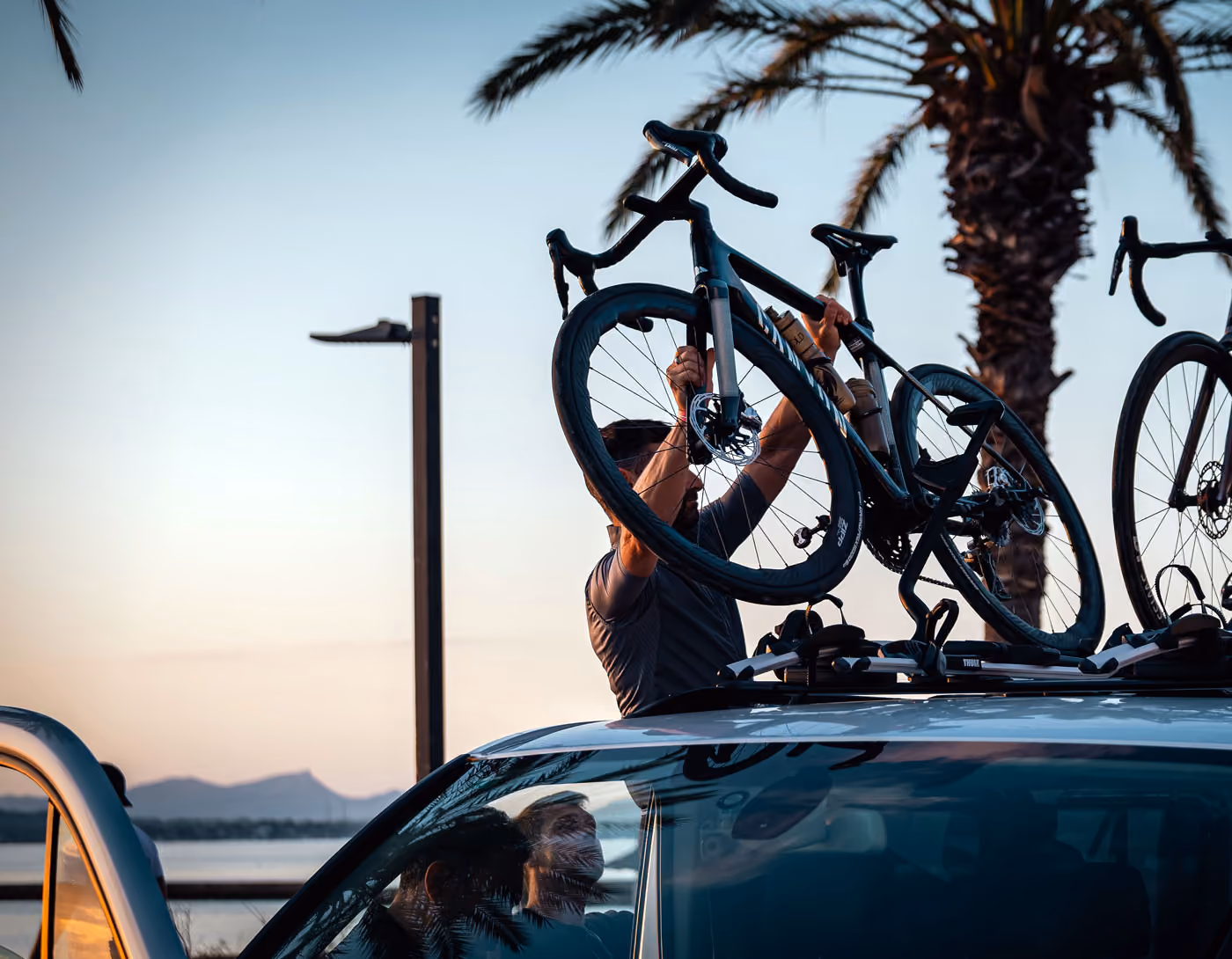 Person securing a black road bike onto the roof rack of a car at sunset near palm trees and water.