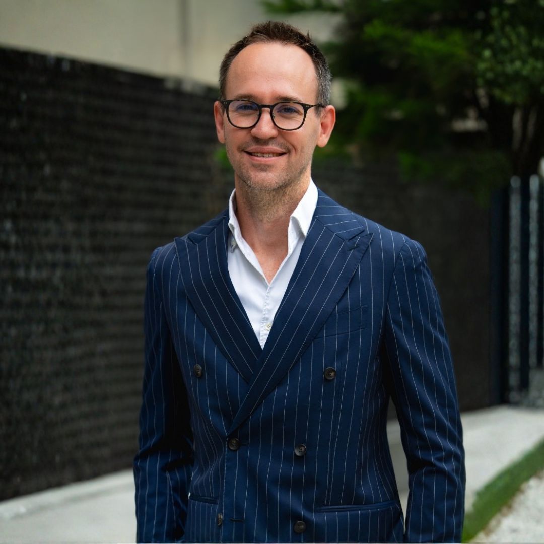 Carl wearing glasses and a dark suit with a white shirt standing in an urban setting with blurred people and glass buildings in the background.
