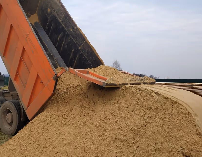 Orange dump truck unloading sand with raised hydraulic bed at construction material yard