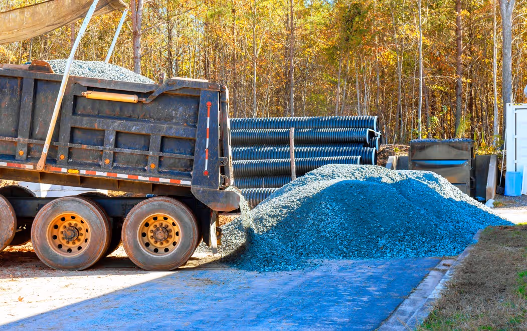Dump truck unloading blue stone gravel near corrugated drainage pipes at construction site