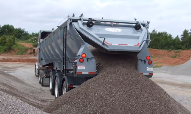 Live bottom trailer unloading aggregate material through rear discharge gate onto construction site roadway