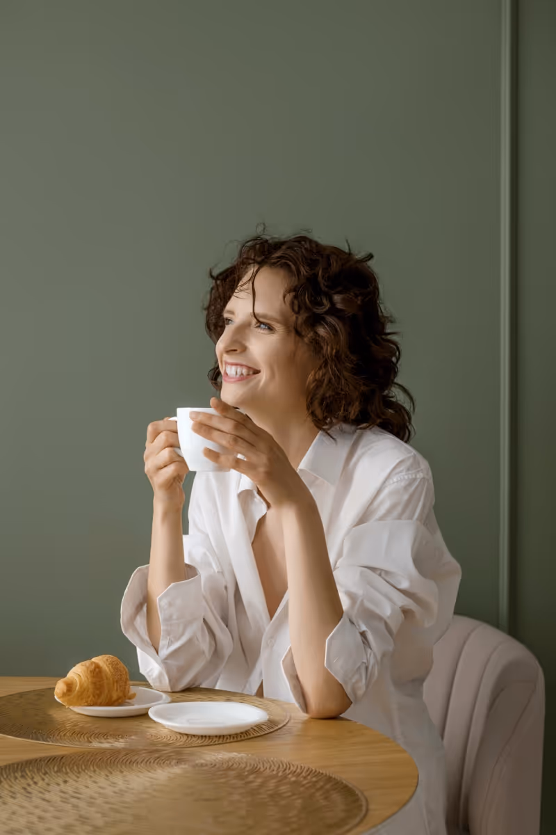Smiling woman with curly hair wearing a white shirt sitting at a table holding a white cup with a croissant on a plate nearby.