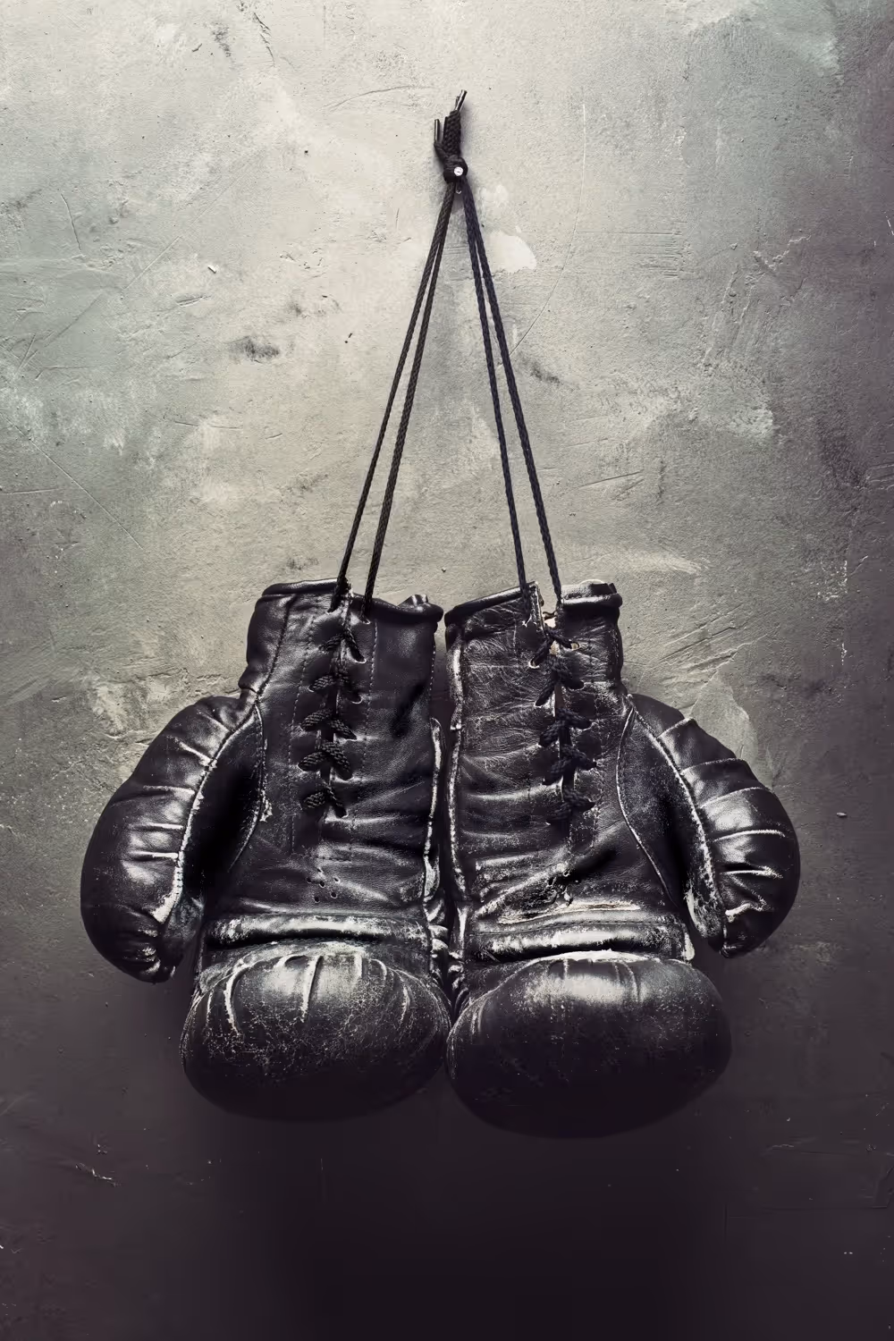 Worn black boxing gloves hanging by their laces on a textured gray wall.