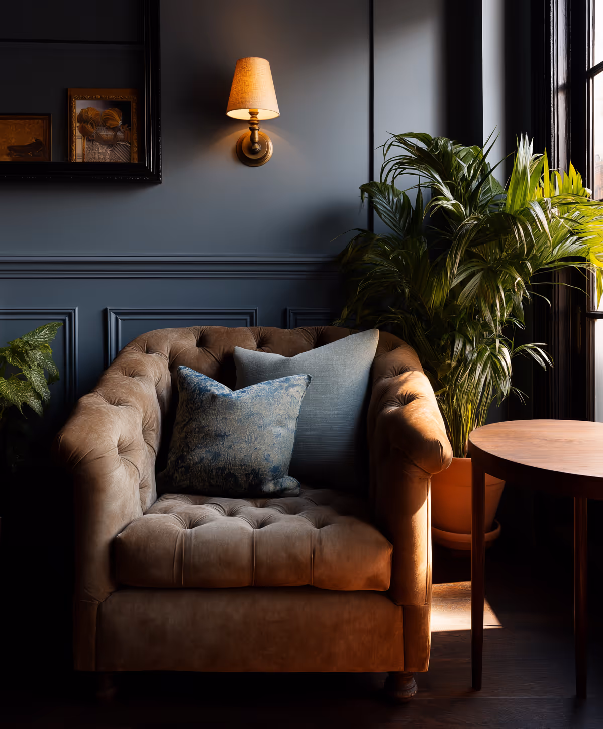 Brown tufted armchair with two decorative pillows next to a potted plant and round wooden table under a lit wall sconce.
