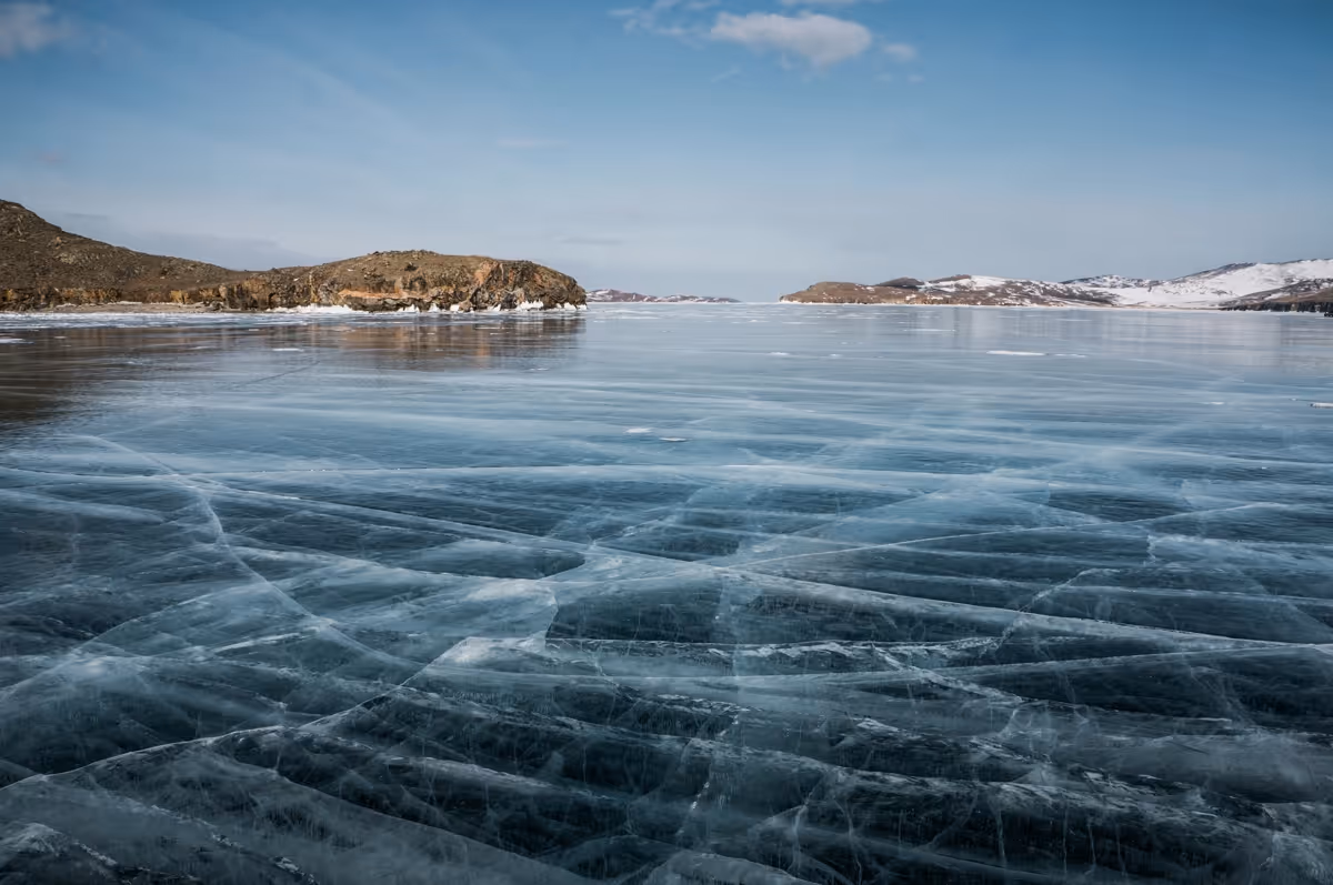 Frozen lake with visible cracks on the ice and rocky snow-dusted hills in the distance under a clear blue sky.