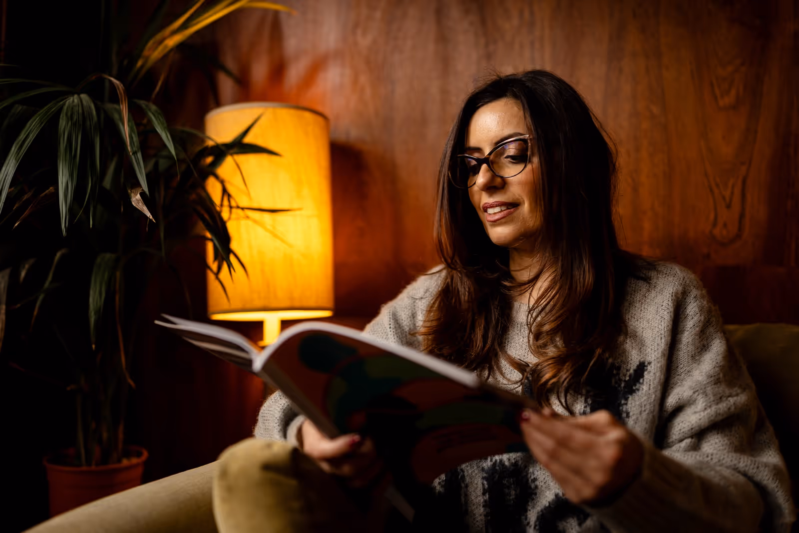Woman with glasses reading a book indoors, sitting on a couch near a lit yellow lamp and a large plant.