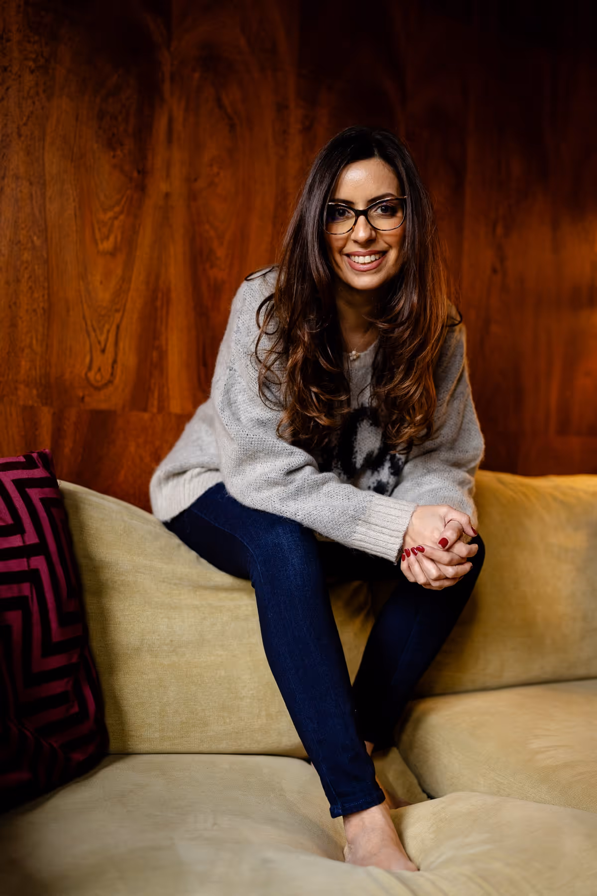 Smiling woman with glasses, wearing a gray sweater and dark jeans, sitting barefoot on a beige couch against a wooden wall.