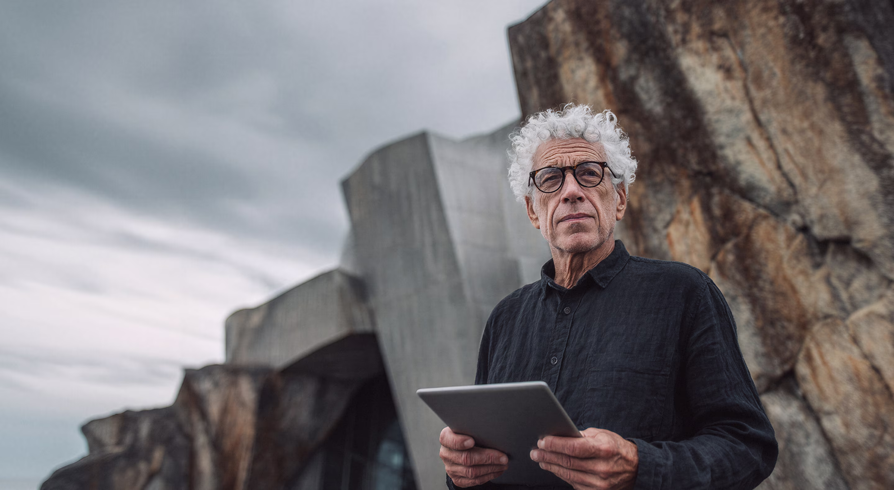 Homme âgé aux cheveux gris bouclés tenant une tablette devant une structure en béton moderne et un rocher.
