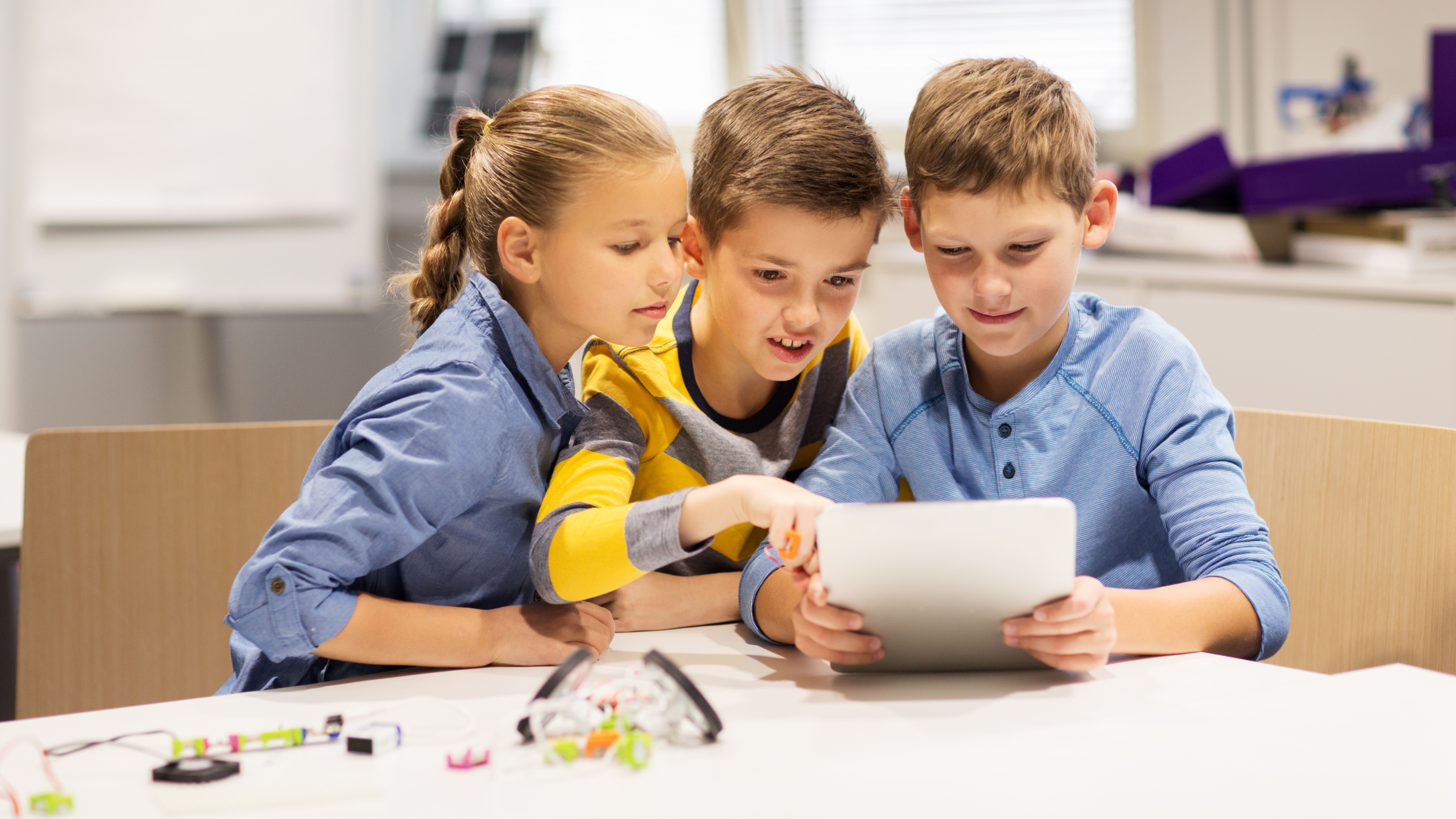 Three children closely looking at a tablet while sitting at a table with electronic components.